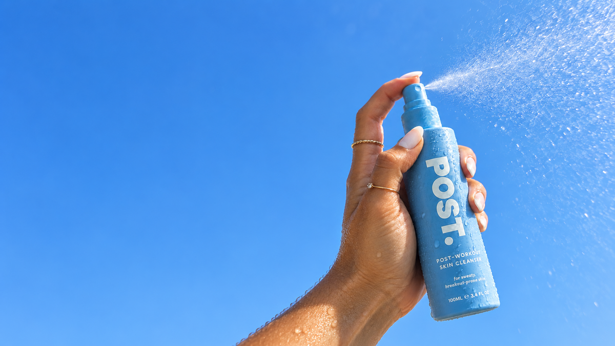 A hand holding a light blue POST. Post-Waterproofing Nail Cleanser spray bottle, spraying water against a bright blue sky.